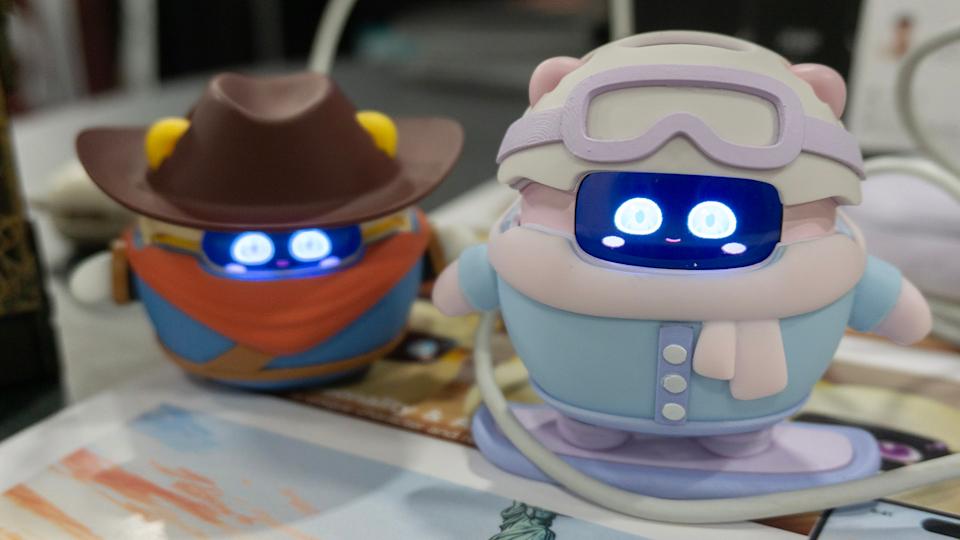 Two Sweekar devices are pictured on a table, one wearing a pink and blue snowboarder outfit and the other (behind it) wearing a cowboy hat and outfit