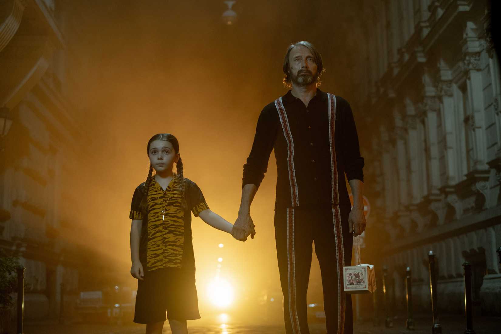 Mads Mikkelsen and Sophie Sloan in Bryan Fuller's Dust Bunny. Sloan wears a tiger-striped shirt and shorts, while Mikkelsen wears a finely decorated shirt and pants. A bright light shines from the alleyway behind them.